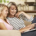 Mother With Teenage Daughter Sitting On Sofa At Home Using Lapto